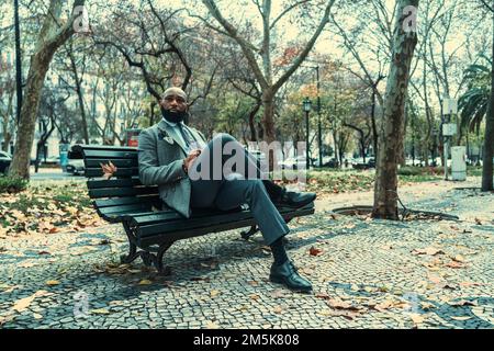 Ein gut aussehender, bärtiger, schwarzer Unternehmer in einem eleganten Outfit, der auf einer hölzernen Straßenbank auf dem Bürgersteig sitzt; ein afroamerikanischer Busin Stockfoto