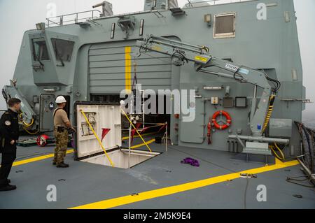 Matrosen an Bord des Patrouillenschiffs HDMS Triton der Königlichen Dänischen Marine heben mit einem Hydraulikkran einen Außenbordmotor aus einem Lagerfach. Stockfoto