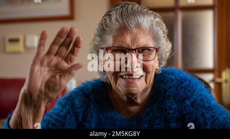 Porträt einer glücklichen, aufgeregten, weißen 90s-Frau, die ein Videogespräch genießt, mit der Hand hallo winkt und im Wohnzimmer auf die Kamera der Webcam schaut. Frohe Seniorin Stockfoto