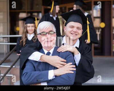 Vater und Sohn umarmen sich bei der Abschlussfeier. Der Elternteil gratuliert dem Hochschulabsolventen. Stockfoto