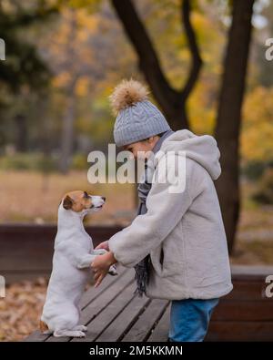 Kaukasisches Mädchen hält einen Hund bei den Pfoten für einen Spaziergang im Herbstpark. Jack Russell Terrier steht auf seinen Hinterbeinen auf einer Bank. Stockfoto