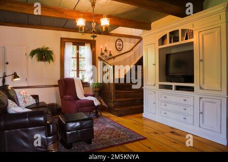 Dunkelbraunes Ledersofa mit Fußhocker und gepolstertem Sessel neben Holztreppe und gebleichter Wandschrank im Wohnzimmer im alten Haus. Stockfoto