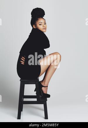 Wunderschön in ihrem kleinen schwarzen Kleid. Studioaufnahme einer modischen jungen Frau, die auf einem Hocker in einem schwarzen Kleid posiert. Stockfoto