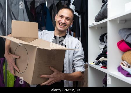 Glücklicher, fröhlicher, männlicher Mann in legerer Kleidung, der eine Pappschachtel trägt und am Umzugstag mit verschwommenen Staffeleien und Kartons in die Kamera schaut Stockfoto