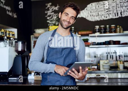 Es ist ein modernes Werkzeug für einen modernen Geschäftsinhaber. Ein gutaussehender junger Barista, der ein Tablet in seinem Café verwendet. Stockfoto