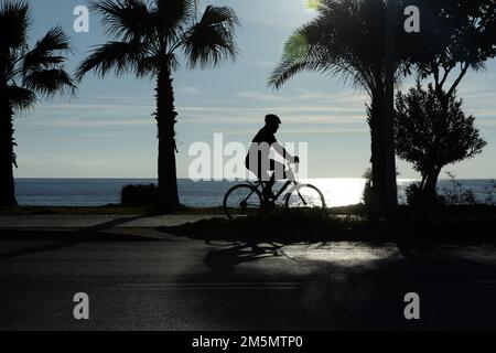 Silhouette eines Mannes, der mit dem Fahrrad vor dem Hintergrund eines Sonnenuntergangs auf dem Meer fährt. Stockfoto