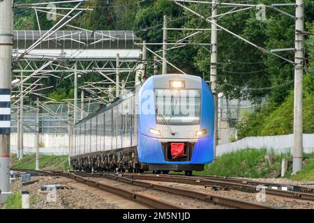 Der blaue Vorstadtzug fährt aus der Ferne mit eingeschalteten Scheinwerfern auf dem Weg zwischen den grünen Bäumen Stockfoto