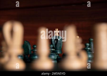 Schwarzer König mit weißem Kreuz im Hintergrund zwischen weißen Schachfiguren, die unscharf sind, mit braunem Holzhintergrund. Konzept: Schach Stockfoto