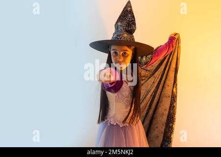 Fröhliches, gruseliges Halloween-Konzept: Kleines Hexenmädchen spielt paranormale Halloween-Rolle und macht einen gruseligen Blick auf gelbem Hintergrund. Kinder erschrecken und machen Stockfoto