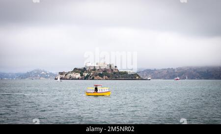 Alcatraz an einem bewölkten Morgen mit einem kleinen gelben Schiff, das in der Mitte des Vordergrunds vorbeifährt. Stockfoto