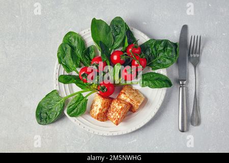 Gebratener Quark Mozzarella halloumi mit grünem Spinat und Kirschtomaten auf einem Zweig auf einem grauen Betontisch Stockfoto