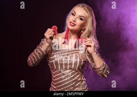 Eine blonde, attraktive Jungfrau mit perfekter Frisur und leuchtendem Make-up, gekleidet in einem goldenen, glänzenden Kleid, posiert mit Spielmarken in ihren Händen. Schlag Zu Stockfoto