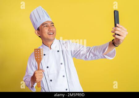 asiatischer Chefkoch trägt ein Selfie mit Kochjacke, während er einen Teigschaber hält Stockfoto