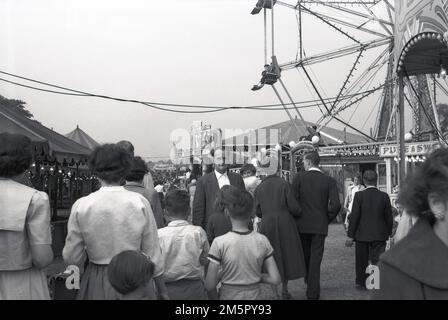 1950er Jahre, historisch, Besucher, die neben den Fahrgeschäften und Ständen auf einem Jahrmarkt spazieren gehen, mit Menschen auf einem Riesenrad, England, Großbritannien. Stockfoto