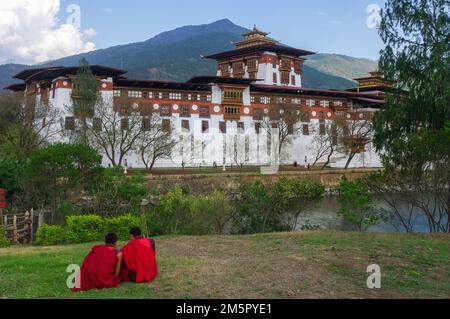 Malerischer Landschaftsblick auf Punakha dzong im Westen von Bhutan, mit anonymen buddhistischen Novizen, die auf dem Gras sitzen Stockfoto