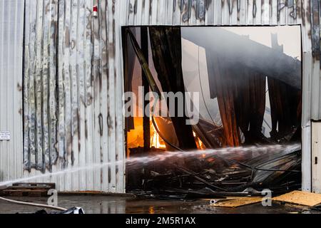 Wehr, Deutschland. 30. Dezember 2022. Flammen können durch das Tor einer ausgebrannten Industriehalle gesehen werden, während im Vordergrund ein Wasserstrahl in das Gebäude spritzt. Ein Brand in mehreren Gebäuden an einem Industriestandort hat Schäden im Wert von mehreren Millionen Euro verursacht. Die Feuerwehr war mit etwa 160 Notfallpersonal vor Ort. Kredit: Philipp von Ditfurth/dpa/Alamy Live News Stockfoto