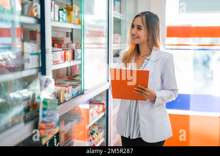 Schöne Apothekerin in Uniform, die mit dem Klemmbrett in der Apotheke arbeitet Stockfoto