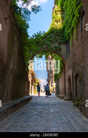 Touristen, die durch einen mit Efeu bedeckten Steinbogen auf der Kopfsteinpflasterstraße spazieren, Civita di Bagnoregio, Viterbo, Latium, Italien Stockfoto