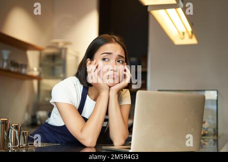 Porträt einer koreanischen Frau, Barista im Café, sah traurig und runzelig aus, grimmige Enttäuschung während der Arbeit im Café, lehnte sich an die Theke, stand Stockfoto