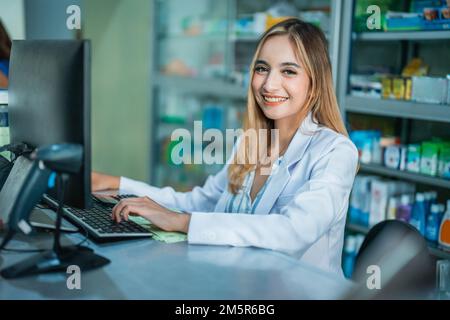 Wunderschöner asiatischer Apotheker in Uniform lächelt bei der Arbeit am Computer Stockfoto