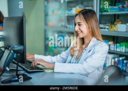 Schöner asiatischer Apotheker in Uniform mit Computer in der Apotheke Stockfoto
