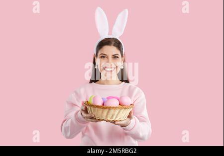 Glückliches Mädchen in pinkfarbenem Sweatshirt mit Hasenohren, das einen Korb mit bunten Eiern auf pinkfarbenem Hintergrund gibt. Stockfoto