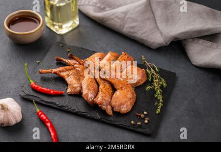 Gegrillte Wachtelbeine in Marinade und Gewürzen auf schwarzem Schiefer mit frischen Kräutern, Öl und Sauce auf dunklem Hintergrund. Gesunde Ernährung. Speicherplatz kopieren. Stockfoto