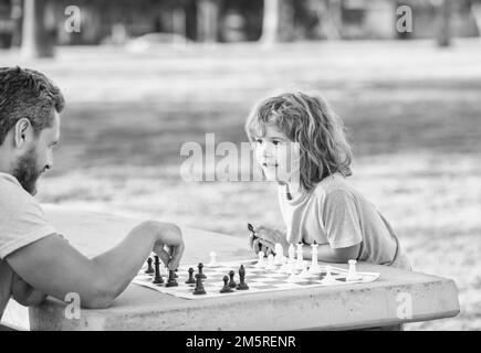 Glückliche Familie von Vater und Sohn Junge spielen Schach auf dem Tisch im Park im Freien, Schachwettbewerb Stockfoto