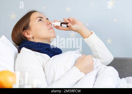 Junge Frau mit schwerer Erkältung, die im Bett unter der Decke liegt und Nasentropfen einnimmt Stockfoto