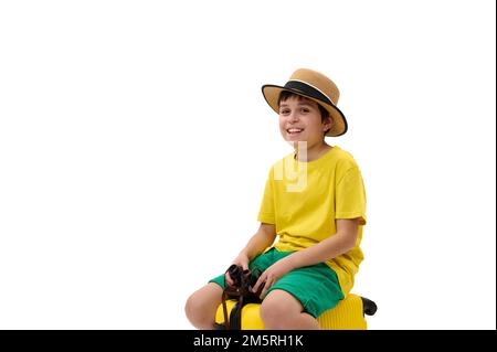 Ein charmanter Teenager mit Strohhut, gelbem T-Shirt, auf dem Weg in die Sommerferien, der auf dem Koffer auf die Kamera schaut Stockfoto