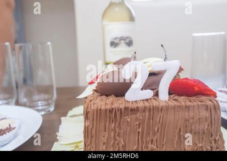 Geburtstagskuchen mit Kerzen in der Form von Nummer 27 auf einem Tisch für ein paar Leute und eine Flasche Wein im Hintergrund, Erwachsenentarif-Konzept Stockfoto
