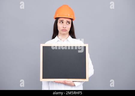 Junge, lustige, traurige Bauleiterin. Frau Baumeister isoliertes Porträt mit Schutzhelm und Brett. Grauer Hintergrund, leerer Bereich, Kopierbereich. Stockfoto