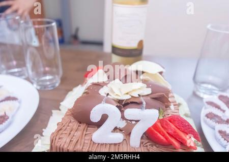 Geburtstagskuchen mit Kerzen in der Form von Nummer 27 auf einem Tisch für ein paar Leute und eine Flasche Wein im Hintergrund, Erwachsenentarif-Konzept Stockfoto