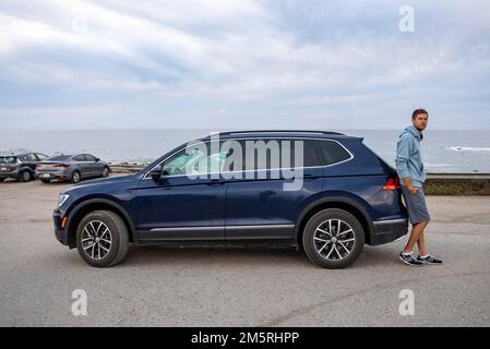 Mann lehnt sich auf blaues Auto mit Meereslandschaft und bewölktem Himmel im Hintergrund Stockfoto