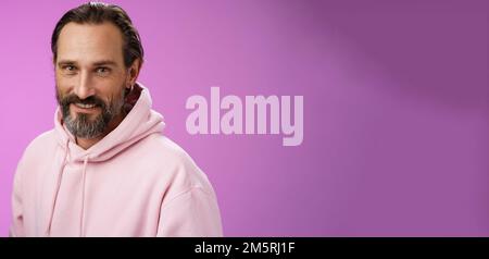 Charming verführerisch gut aussehend bärtig stilvolle Erwachsene männliche Modell Ohrring rosa hoodie Smiling delighted Express Confidence Positivity Fühlen Sie sich glücklich amüsiert Stockfoto