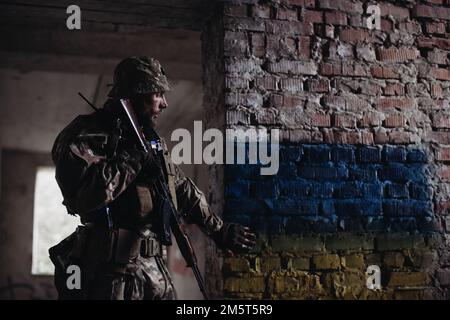 Ein ukrainischer Soldat mit einer Waffe in der Hand steht vor dem Hintergrund der ukrainischen Flagge. Die ukrainische Flagge ist auf einem Ziegelstein gemalt Stockfoto