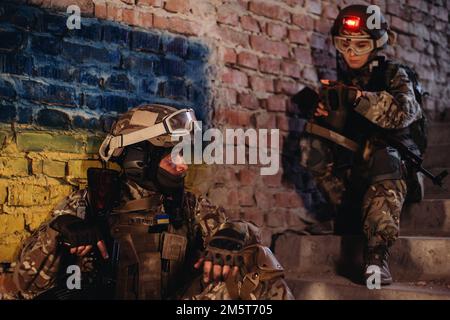 Waffenbrüder, die vor dem Hintergrund der ukrainischen Flagge ruhten. Das ukrainische Militär im einstürzten Gebäude, das sich aus dem Krieg erholt hat. Stockfoto