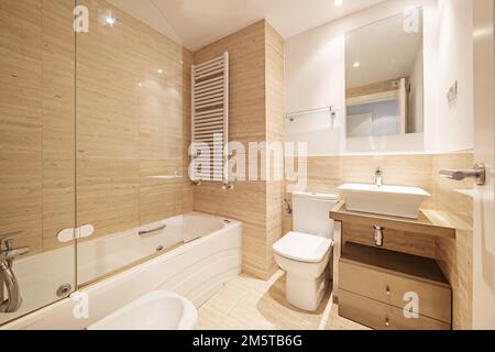 Badezimmer in konventionellem Design mit Marmorfliesen an Wänden und Böden, rechteckigem Spiegel, Schrank mit Schubladen und einem kleinen weißen Porzellanwaschbecken mit Koch Stockfoto