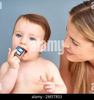 Ich Frage mich, ob es andere Geschmacksrichtungen gibt. Ein bezauberndes Mädchen, das in einen Holzblock beißt, während ihre Mutter zusieht. Stockfoto