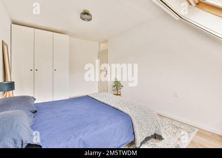 Ein Schlafzimmer mit weißen Wänden und blauem Bett im ganzen Zimmer, es gibt eine kleine Pflanze in der Ecke Stockfoto