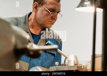 Das Porträt eines 50-55 Jahre alten Turners mit Brille bei der Arbeit. Ältere Metallwender arbeiten in der Werkstatt an der Maschine hinter der Arbeit. Stockfoto