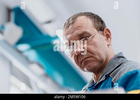 Porträt einer 50-55 Jahre alten Turner mit Brille bei der Arbeit. Bottom-up-Ansicht. Ältere Metallwender arbeiten in der Werkstatt an der Maschine hinter der Arbeit. Stockfoto