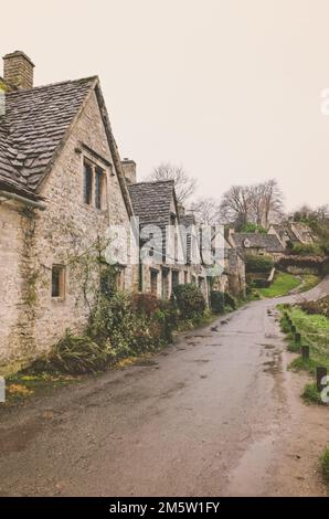 Ein vertikales Bild einer nassen Straße entlang einer Reihe historischer, typischer Cotswold-Hütten in Bibury, England Stockfoto