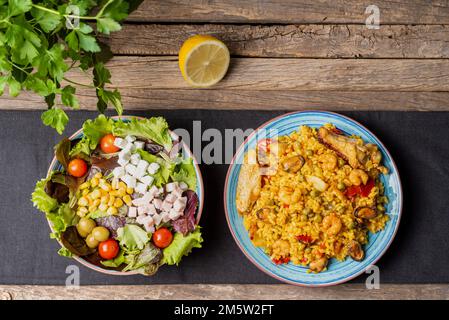 Blauer Teller mit gemischter Paella, Fleisch und Fisch auf einer grauen Tischdecke, begleitet von einer Schüssel mit Gemüse- und Käsesalat auf einem rustikalen Holztisch. Stockfoto