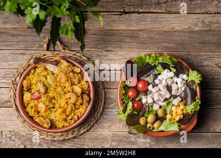Rustikaler Teller mit gemischter Paella, Fleisch und Fisch, begleitet von einer Schüssel mit Gemüse- und Käsesalat, auf einem alten Holztisch. Stockfoto