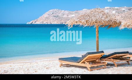 Mykonos Strand im Sommer mit Sonnenschirm und luxuriösen Liegestühlen, blaues Meer mit einem Berg am Elia Beach Mikonos Greece Beach Stockfoto