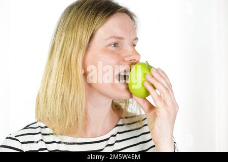 Junge Frau mit Zahnspange, die grünen Apfel beißt, isolierter heller Hintergrund. Studio. Süßes, hübsches Mädchen mit Metallspange auf weißen, klaren Zähnen, das mich lächelt Stockfoto