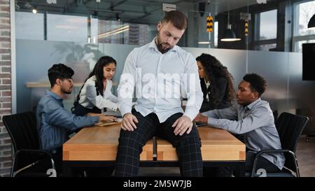 Weißer Geschäftsmann fauler Arbeiter, der mit geschlossenen Augen auf dem Tisch sitzt, tut so, als schliefe er müde, erschöpft vor dem Hintergrund eines multiethnischen Teams Stockfoto