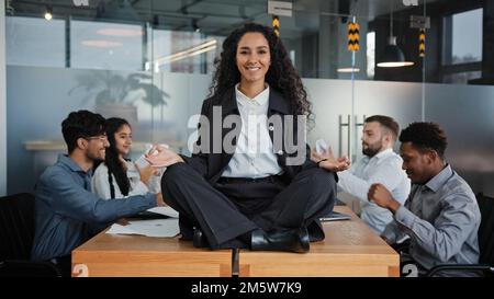 Lächelnde Achtsamkeit Geschäftsfrau glückliche Anführerin, die an einem Tisch in Lotusposition sitzt und in einem Büro meditiert, entspannen im Meditationshintergrund Stockfoto
