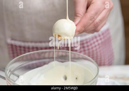 Cake Pops machen: Die Hand hält einen Cake Pops an einem Stock und taucht in eine Schüssel in eine weiße Beschichtung. Stockfoto